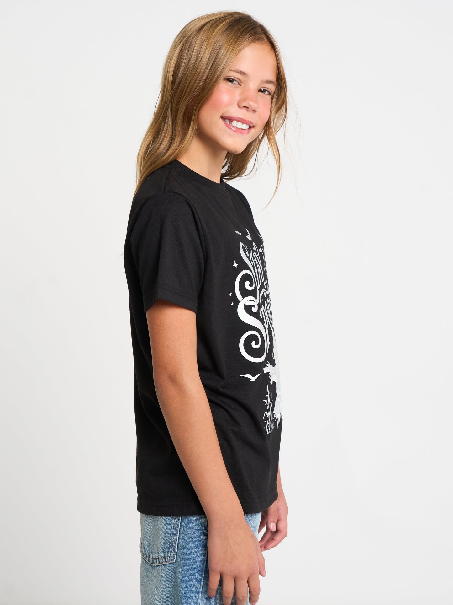 A young girl with blonde hair is shown in profile wearing a black short-sleeve t-shirt with a white graphic print on the front, paired with blue jeans, against a plain white studio background.