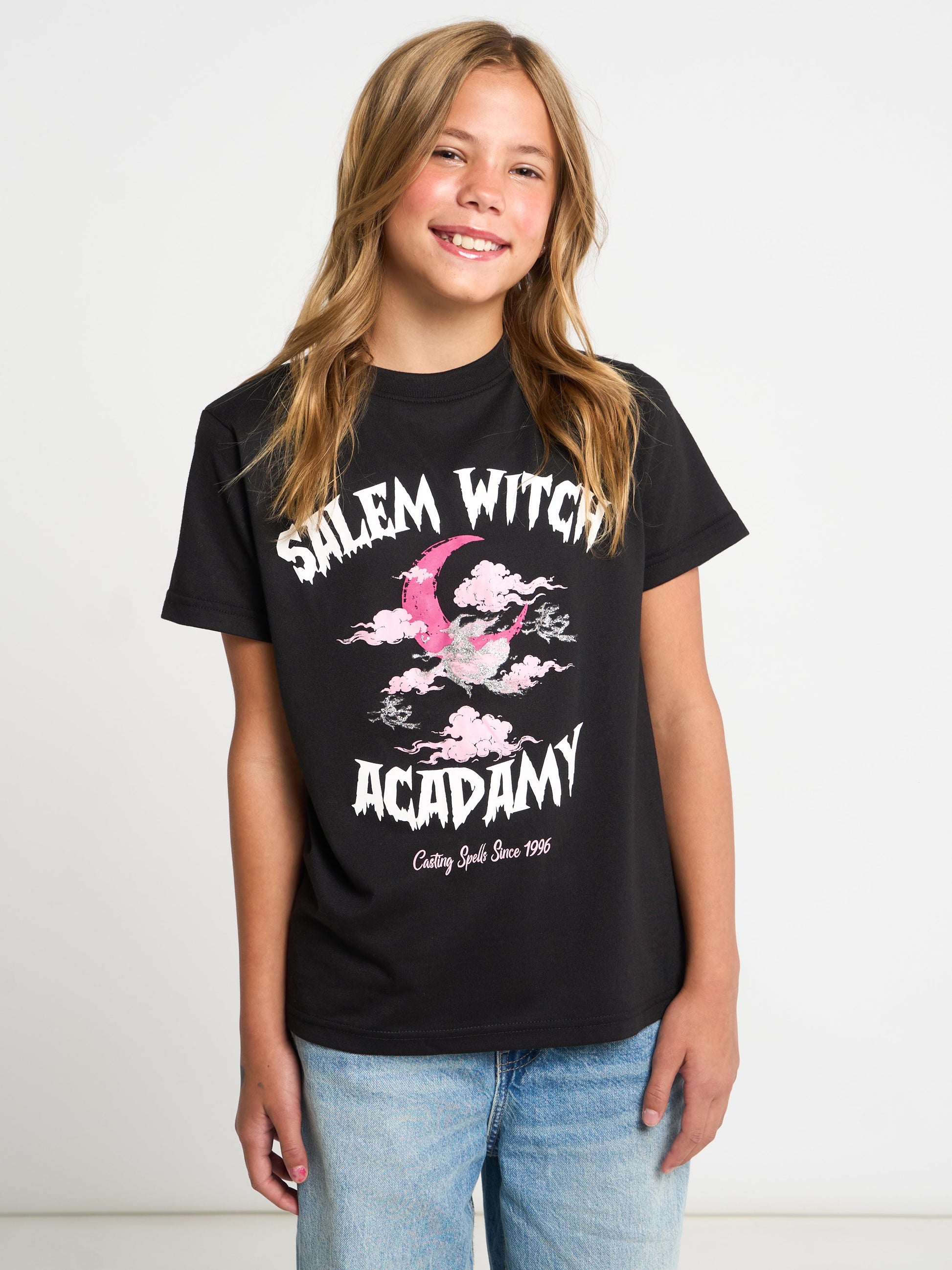 A young girl with blonde hair smiling, wearing a black graphic t-shirt that reads 'Salem Witch Academy' with a pink witch hat illustration, paired with light blue jeans, against a plain white studio background.