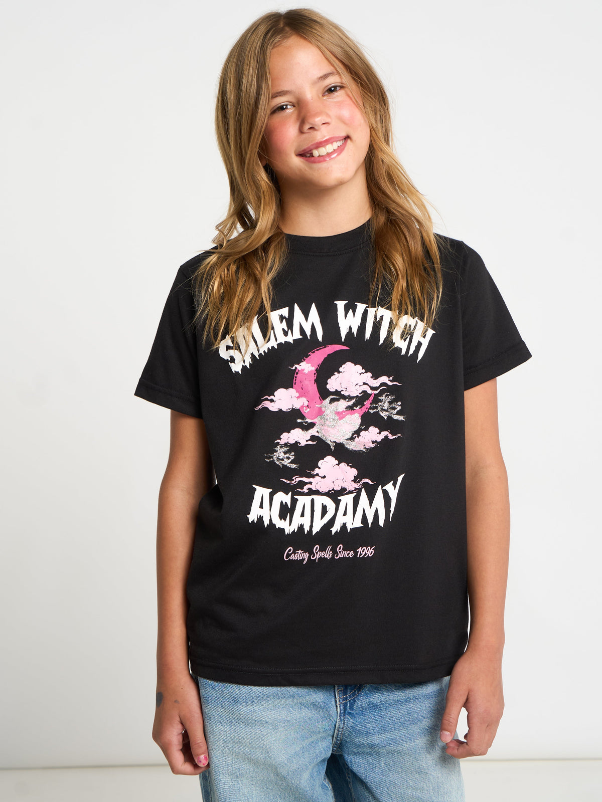 A young girl with long light brown hair smiling, wearing a black graphic t-shirt that reads 'Salem Witch Academy' in white with a pink witch hat illustration, paired with blue jeans, against a plain white background.