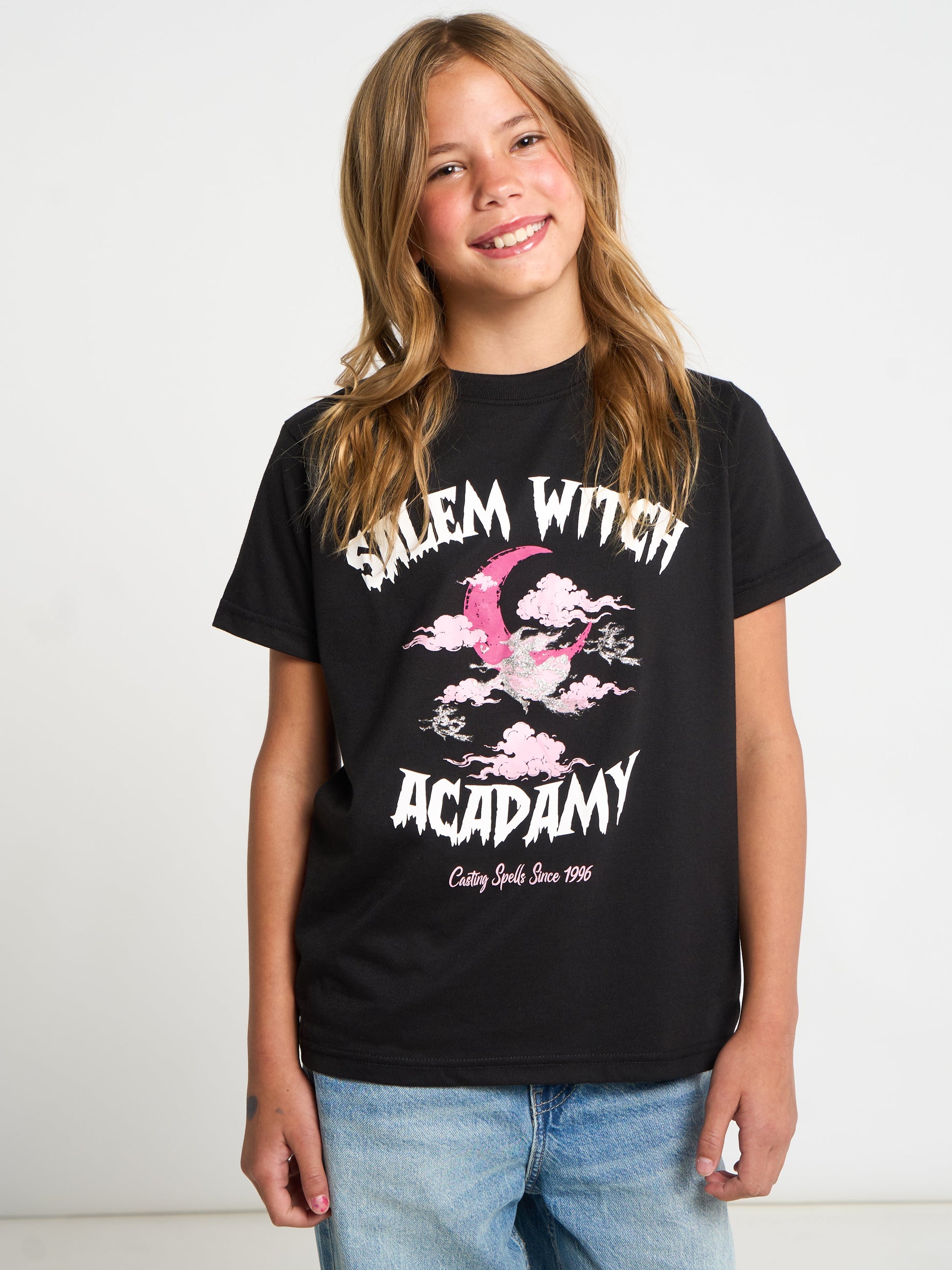 A young girl with long light brown hair smiling, wearing a black graphic t-shirt that reads 'Salem Witch Academy' in white with a pink witch hat illustration, paired with blue jeans, against a plain white background.