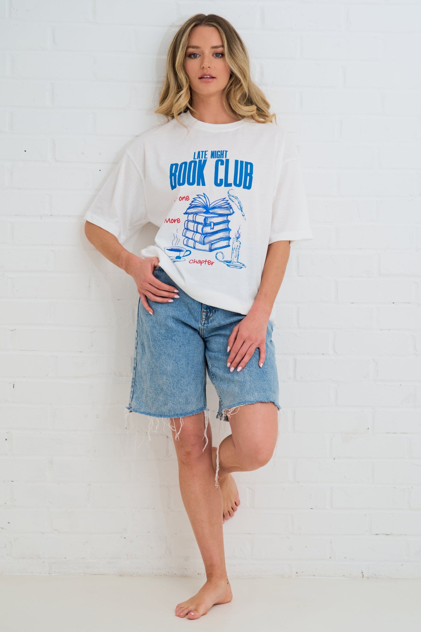 Woman wearing a white t-shirt with a graphic design and denim shorts against a white brick wall.