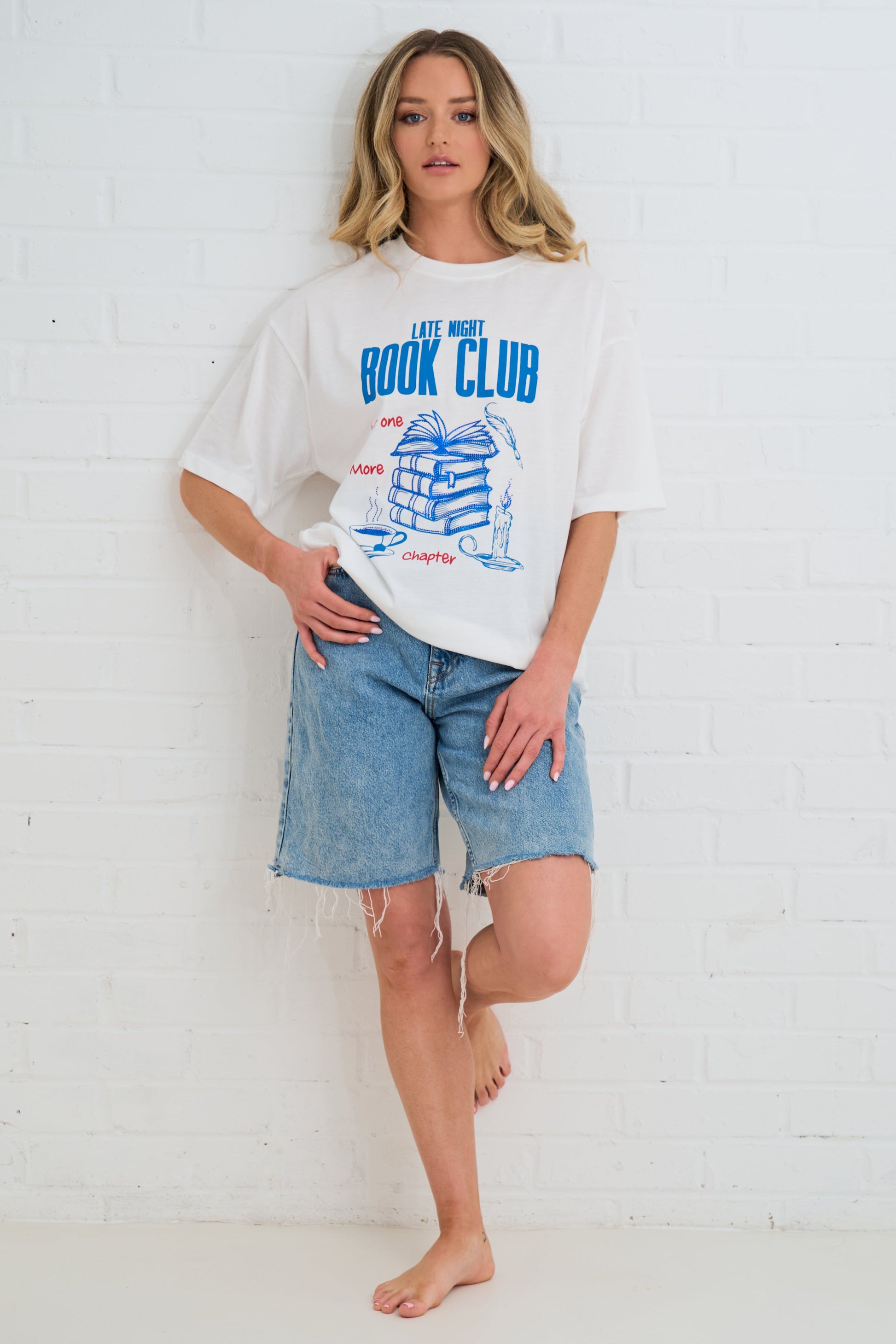 Woman wearing a white t-shirt with a graphic design and denim shorts against a white brick wall.