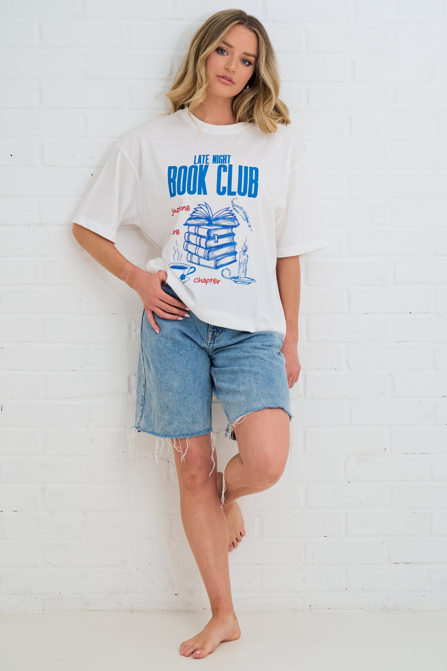 Woman wearing a white t-shirt with 'Book Club' design and denim shorts against a white wall.