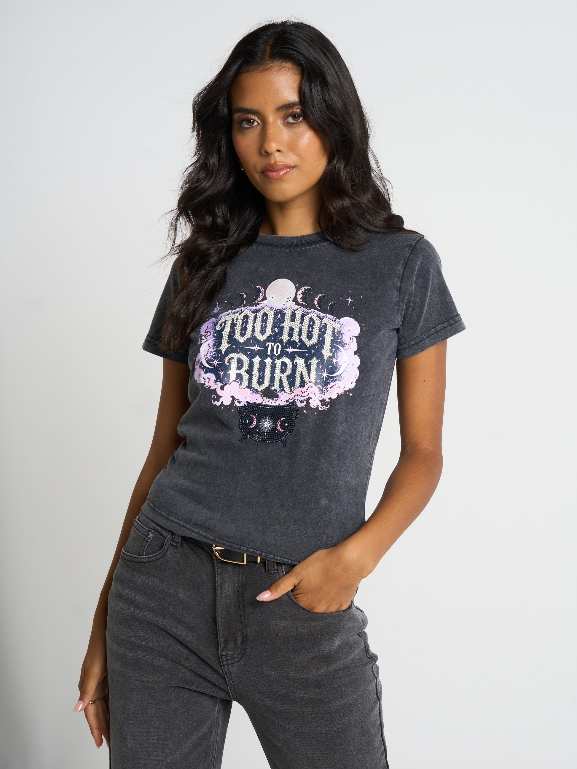 A woman modeling a dark gray acid-wash graphic t-shirt with the text 'TOO HOT TO BURN' in a purple and white celestial design, paired with dark gray jeans against a light gray studio background.