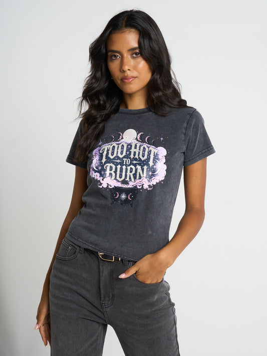 A woman modeling a dark gray acid-wash graphic t-shirt with the text 'TOO HOT TO BURN' in a purple and white celestial design, paired with dark gray jeans against a light gray studio background.