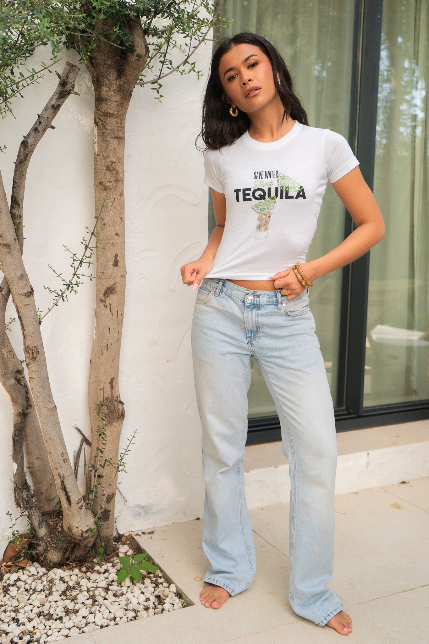 Woman wearing a white t-shirt with 'Life is better with Tequila' graphic and light blue jeans, standing outdoors.