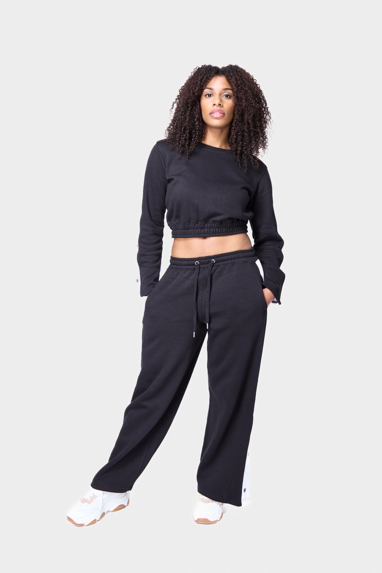 Woman wearing black cropped track suit with white snap-button detailing on the sleeves and pants.