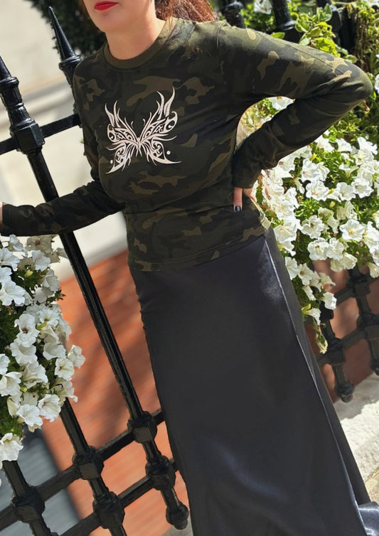 Person wearing a camouflage long-sleeve shirt with a white butterfly design, standing in front of a black metal fence and white flowers.