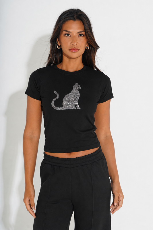A woman modeling a black short-sleeve crop top with a sparkling silver cat graphic on the front, paired with black sweatpants, against a plain white studio background.